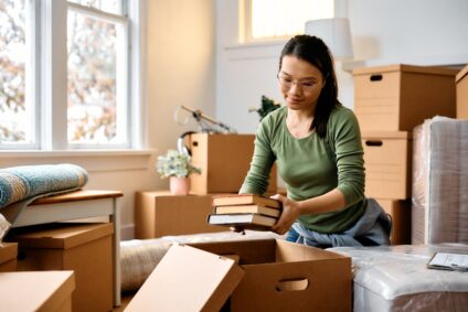 A young woman packs books into a cardboard moving box.