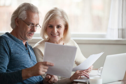 Two older people are looking at a computer and studying papers and discussing a decision.