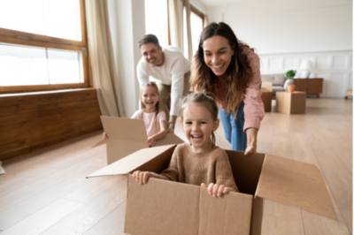 Family laughing while parents push their kids along floor while kids are inside cardboard boxes.