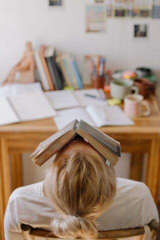 A person leaning back in their desk chair with a book covering their face.