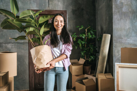 young woman with ficus plant and boxes moving into new house