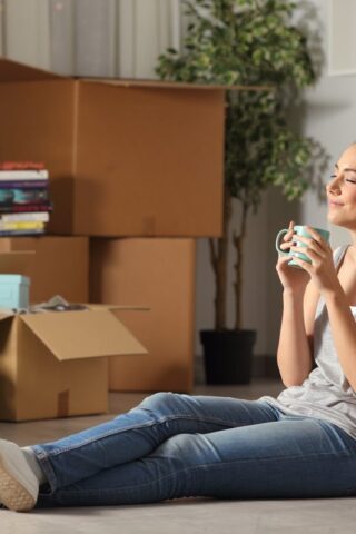 A person sitting on the ground, leaning against a box and smiling while they sip their coffee.