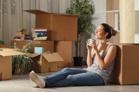 A person sitting on the ground, leaning against a box and smiling while they sip their coffee.