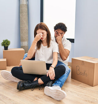 A young, distressed couple sits on the floor surrounded by moving boxes.