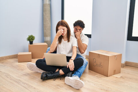 A young, distressed couple sits on the floor surrounded by moving boxes.