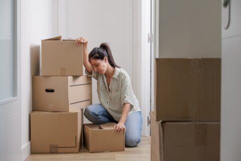 A woman looks tired and frustrated as her head leans against her arm and she kneels among her moving boxes.