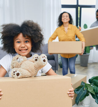 A young girl smiles while holding a moving box with a teddy bear on top. Her parents are holding boxes and smiling in the background.