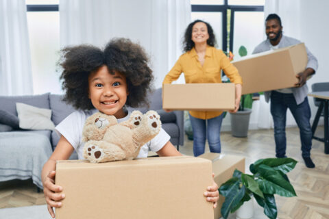 A young girl smiles while holding a moving box with a teddy bear on top. Her parents are holding boxes and smiling in the background.