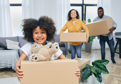 A young girl smiles while holding a moving box with a teddy bear on top. Her parents are holding boxes and smiling in the background.