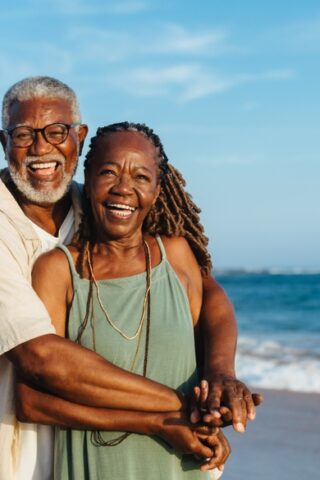 A smiling couple hugs while on a beach with the ocean in the background.