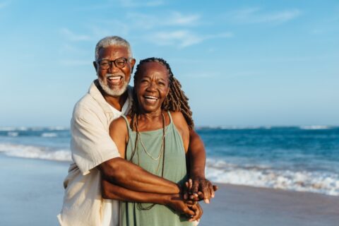 A smiling couple hugs while on a beach with the ocean in the background.
