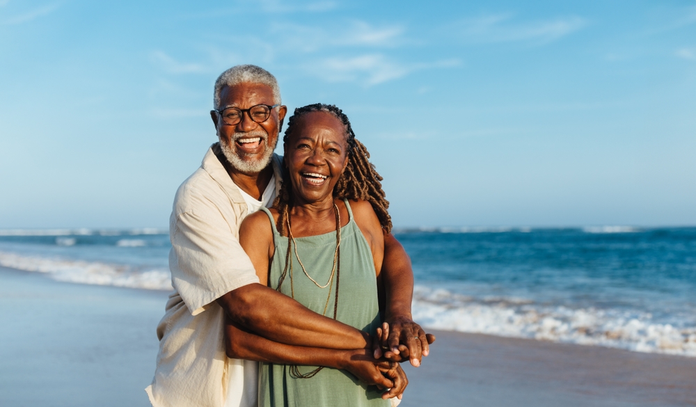 A smiling couple hugs while on a beach with the ocean in the background.