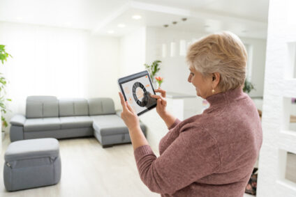 An older woman stands in her living room and uses her tablet to adjust her lights.