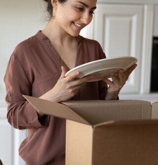 A woman smiles as she packs plates into a cardboard box.