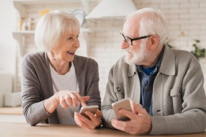 Two older adults look at each other and smile while standing in their kitchen and using their smartphones.