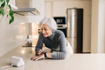 An older woman leans over her kitchen countertop and uses a video calling device.