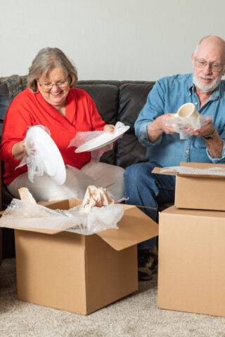 An older adult couple packs cardboard moving boxes in their living room.
