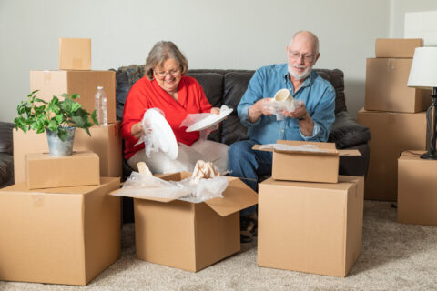 An older adult couple packs cardboard moving boxes in their living room.