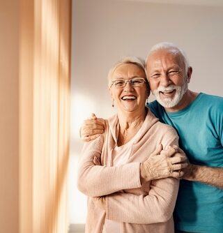 Two older adults hug each other and smile in a house with blank walls.