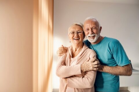 Two older adults hug each other and smile in a house with blank walls.