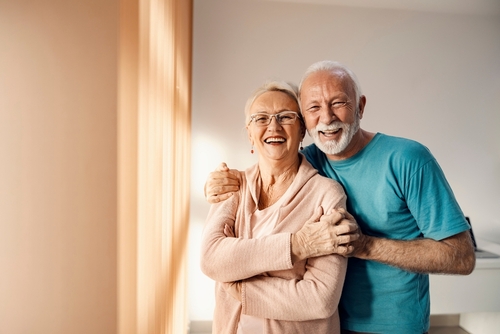 Two older adults hug each other and smile in a house with blank walls.