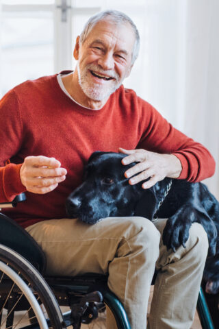An older man in a wheelchair smiles as he pets his dog.
