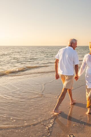 An older adult couple walks on the beach at sunset while holding hands.