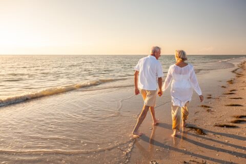 An older adult couple walks on the beach at sunset while holding hands.