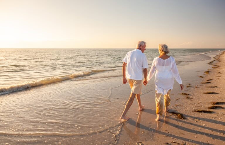 An older adult couple walks on the beach at sunset while holding hands.