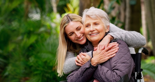 A woman hugs an older woman in a wheelchair from behind.