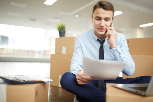 A business man speaks on the phone while surrounded by cardboard boxes in an office.