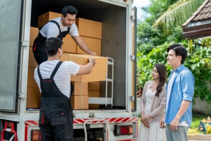 A young couple stands in front of a moving truck as a mover loads cardboard boxes into it.
