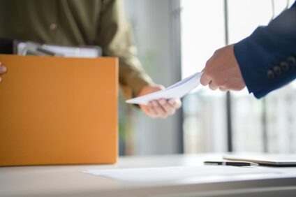 A boss hands an employee a piece of paper over a desk with a cardboard box