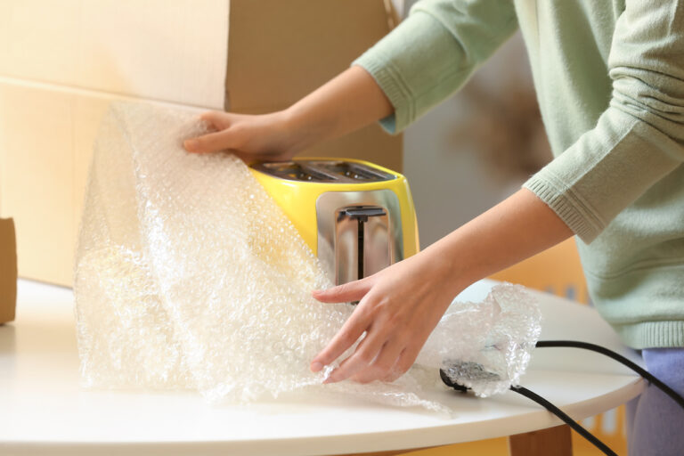A woman wraps a yellow toaster in bubble wrap.