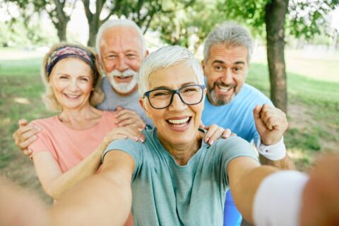 A group of older adults smiles for a selfie while standing in a park.