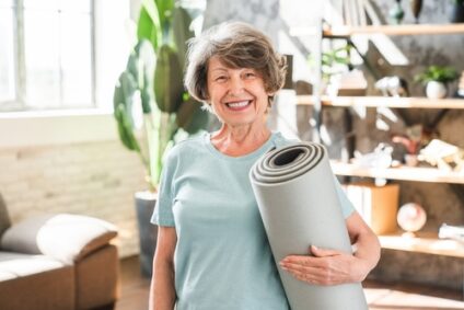 An older woman smiles while holding a yoga mat.